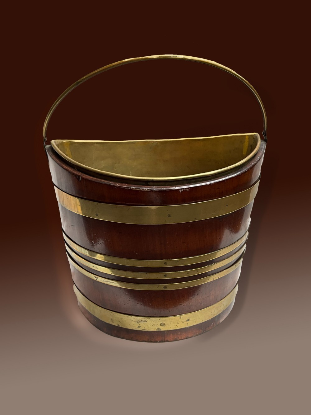 An early 19th-century elliptical mahogany bucket featuring horizontal brass banding, a brass swing handle, and a removable internal brass liner.