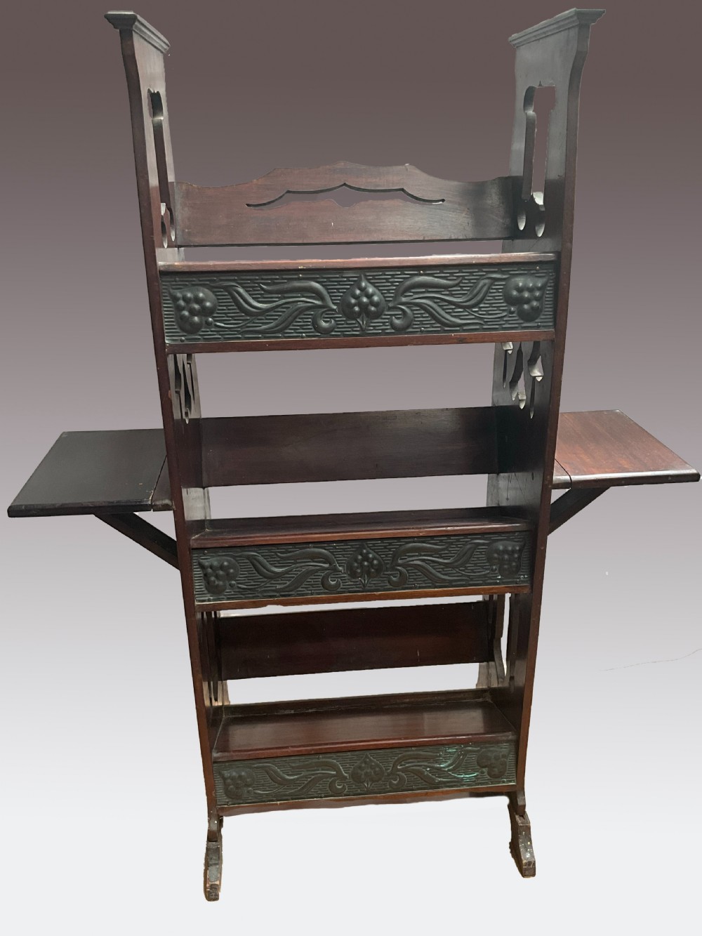 An Art Nouveau dark wood open bookcase featuring side drop-leaf shelves and decorative embossed metal panels with organic motifs.