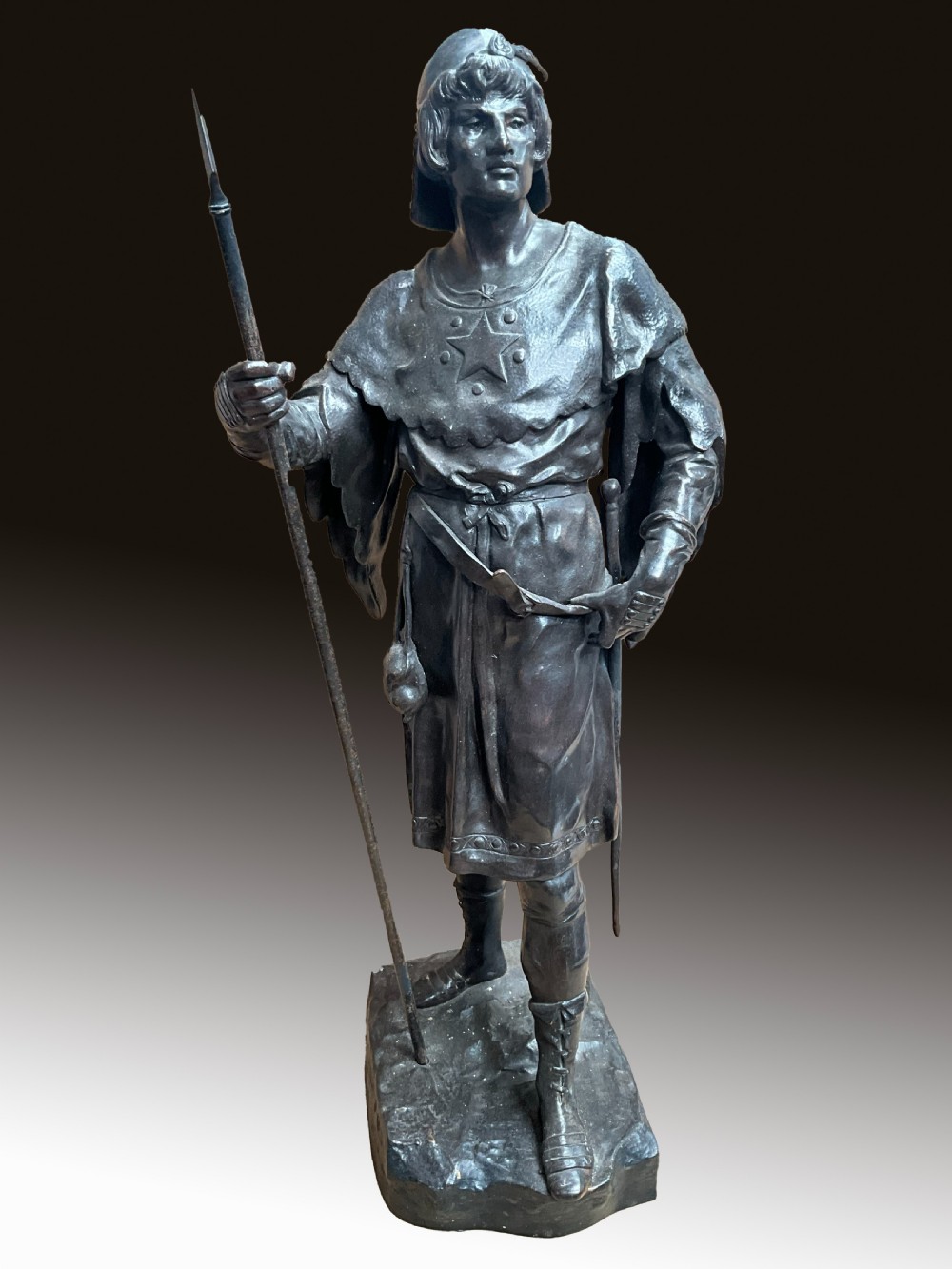 A dark patinated bronze statue of a soldier holding a long spear on a rocky base.