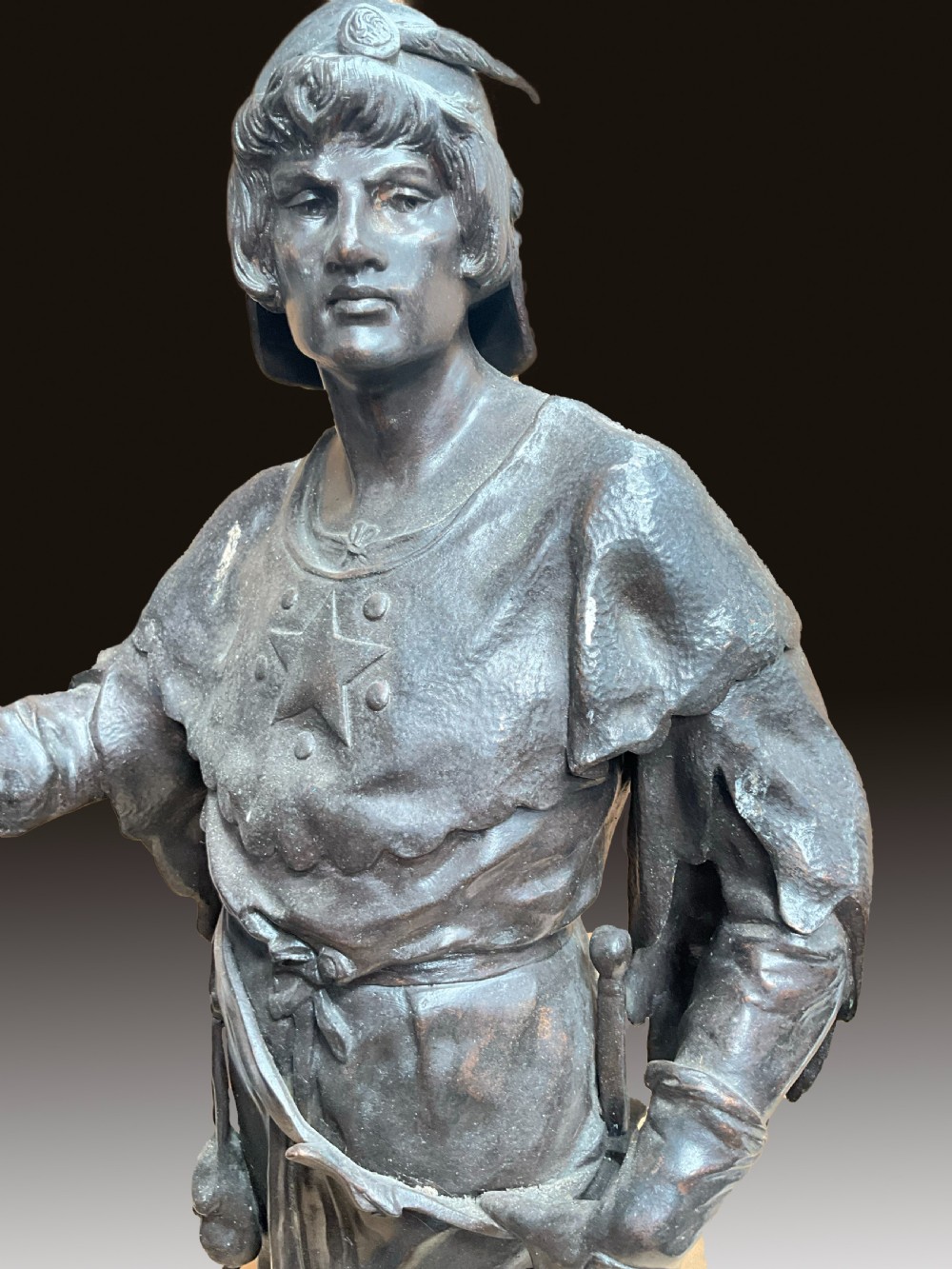 A dark patinated bronze statue of a soldier holding a long spear on a rocky base. View IV