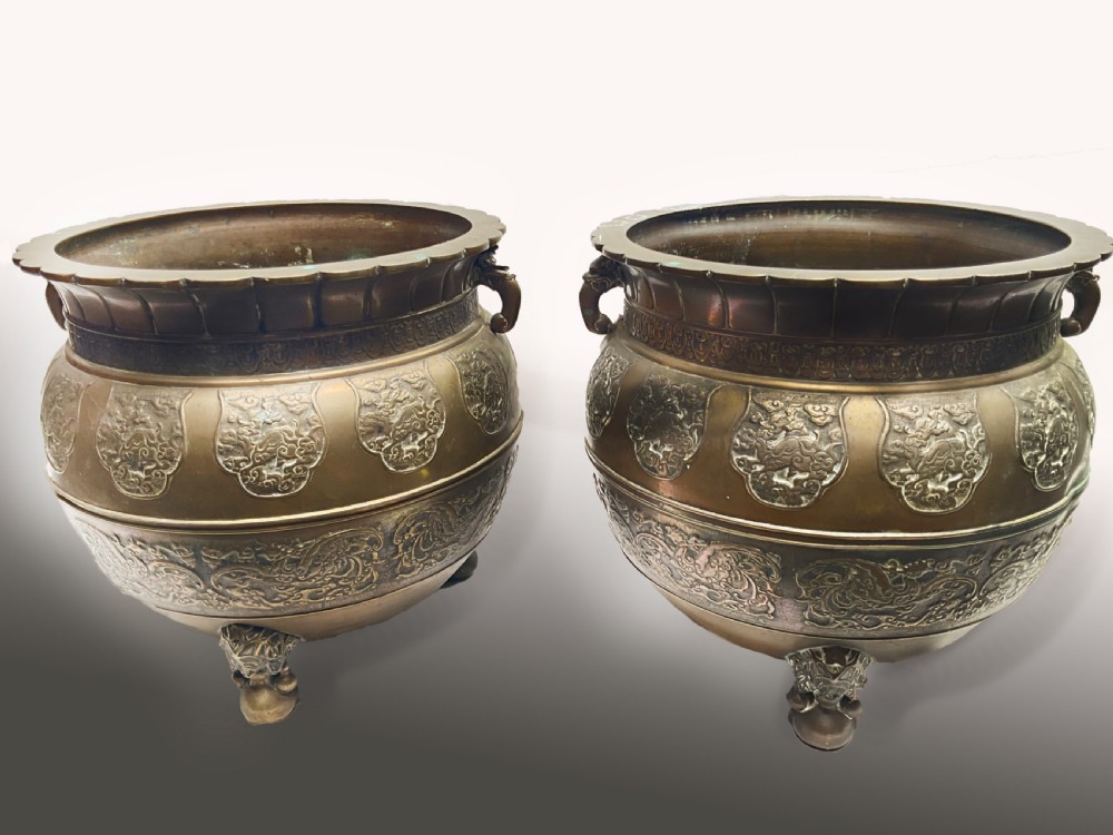 A pair of patinated bronze Oriental jardinieres featuring relief dragon carvings, scalloped rims, and tripod mask feet.