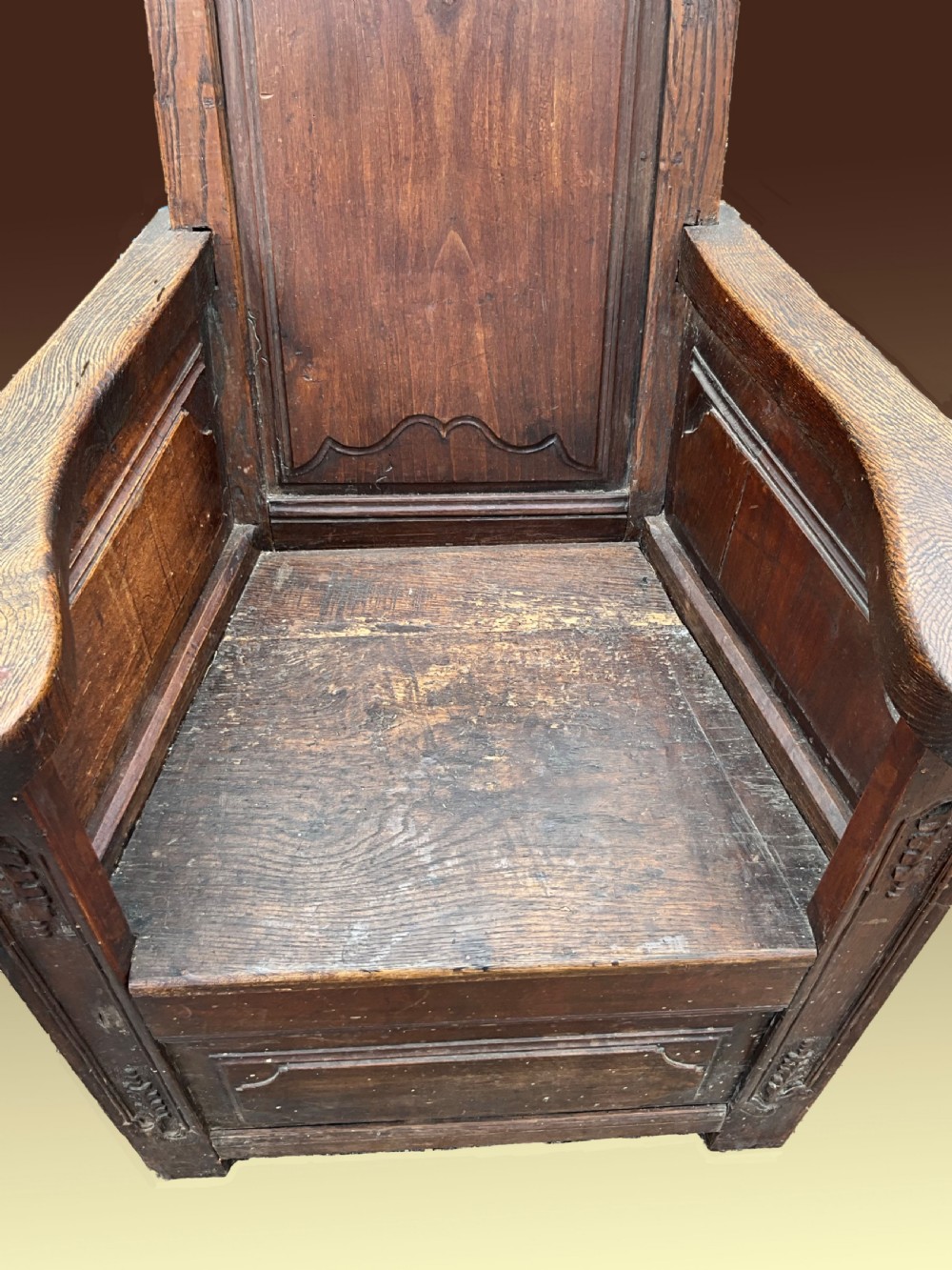 An antique dark oak Dutch country lambing chair featuring a high panelled back, broad flat armrests, and carved vertical accents. View III