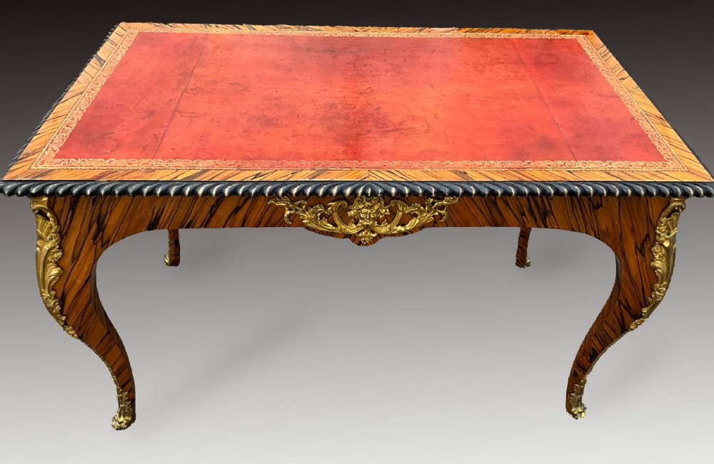 A French zebrawood writing table featuring a red leather top with gilt tooling, curved cabriole legs, and ornate gold-coloured mounts.