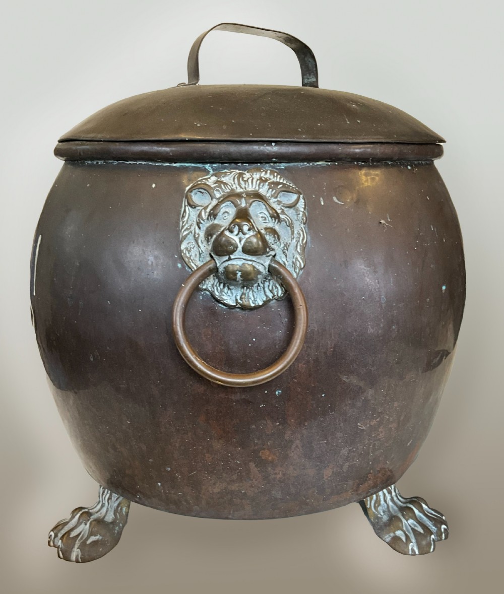 An antique dark copper cauldron featuring a domed lid, top handle, brass lion head mask ring handles, and cast feet.