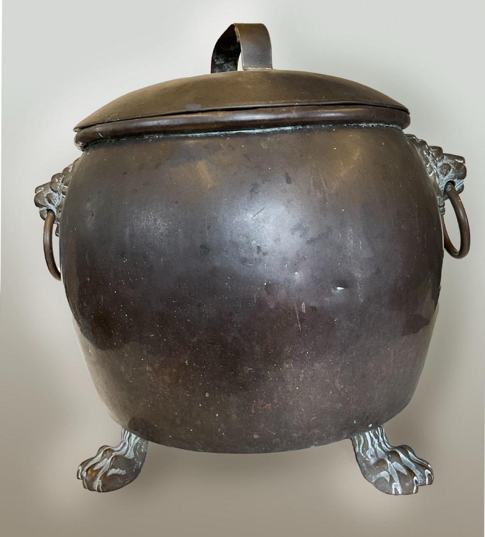 An antique dark copper cauldron featuring a domed lid, top handle, brass lion head mask ring handles, and cast feet. View V