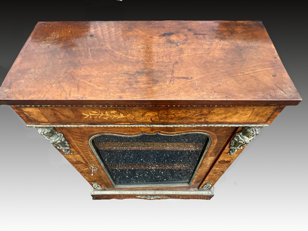 A Victorian burr walnut glazed cabinet featuring floral marquetry inlays, metal mounts, and a single glass door. View VI