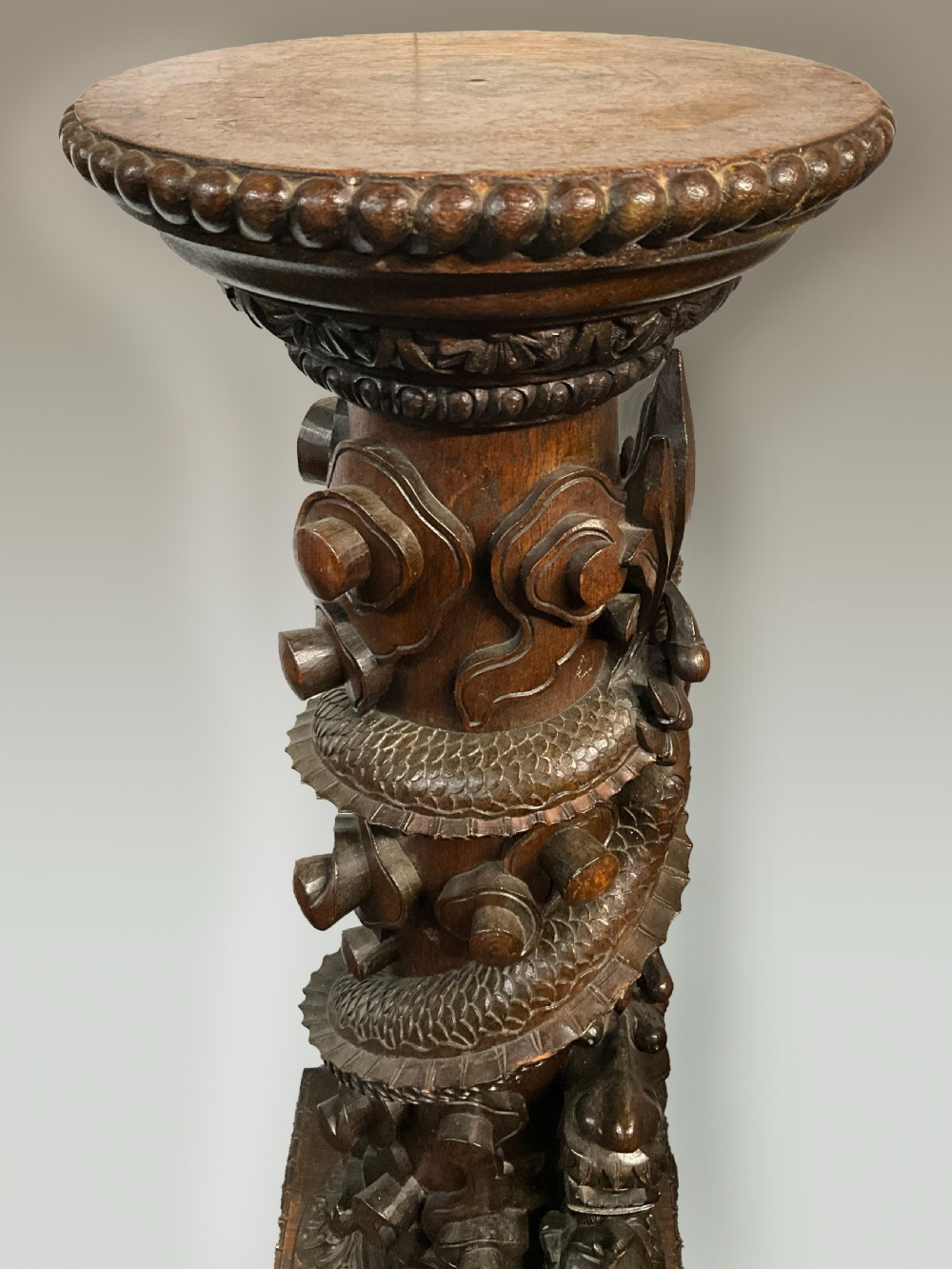 A tall dark brown hand-carved wooden column stand depicting a coiled dragon wrapped around the central shaft above a patterned square base. View III