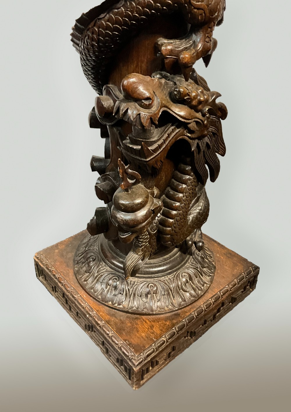 A tall dark brown hand-carved wooden column stand depicting a coiled dragon wrapped around the central shaft above a patterned square base. View V