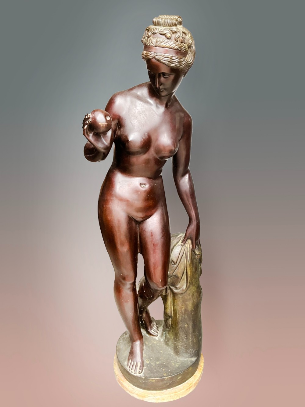 A classical bronze sculpture of a standing female figure holding a small sphere beside draped fabric on a marble base.