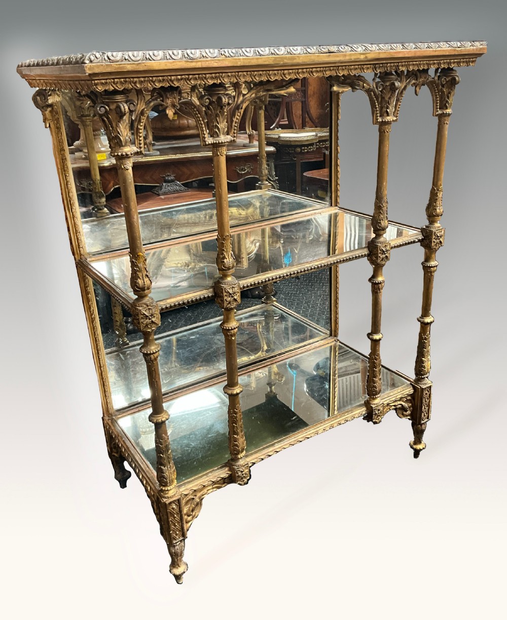 An ornate Victorian giltwood display shelf featuring three glass-tiered levels, a mirrored back panel, and intricate carved gesso columns with decorative moulding.