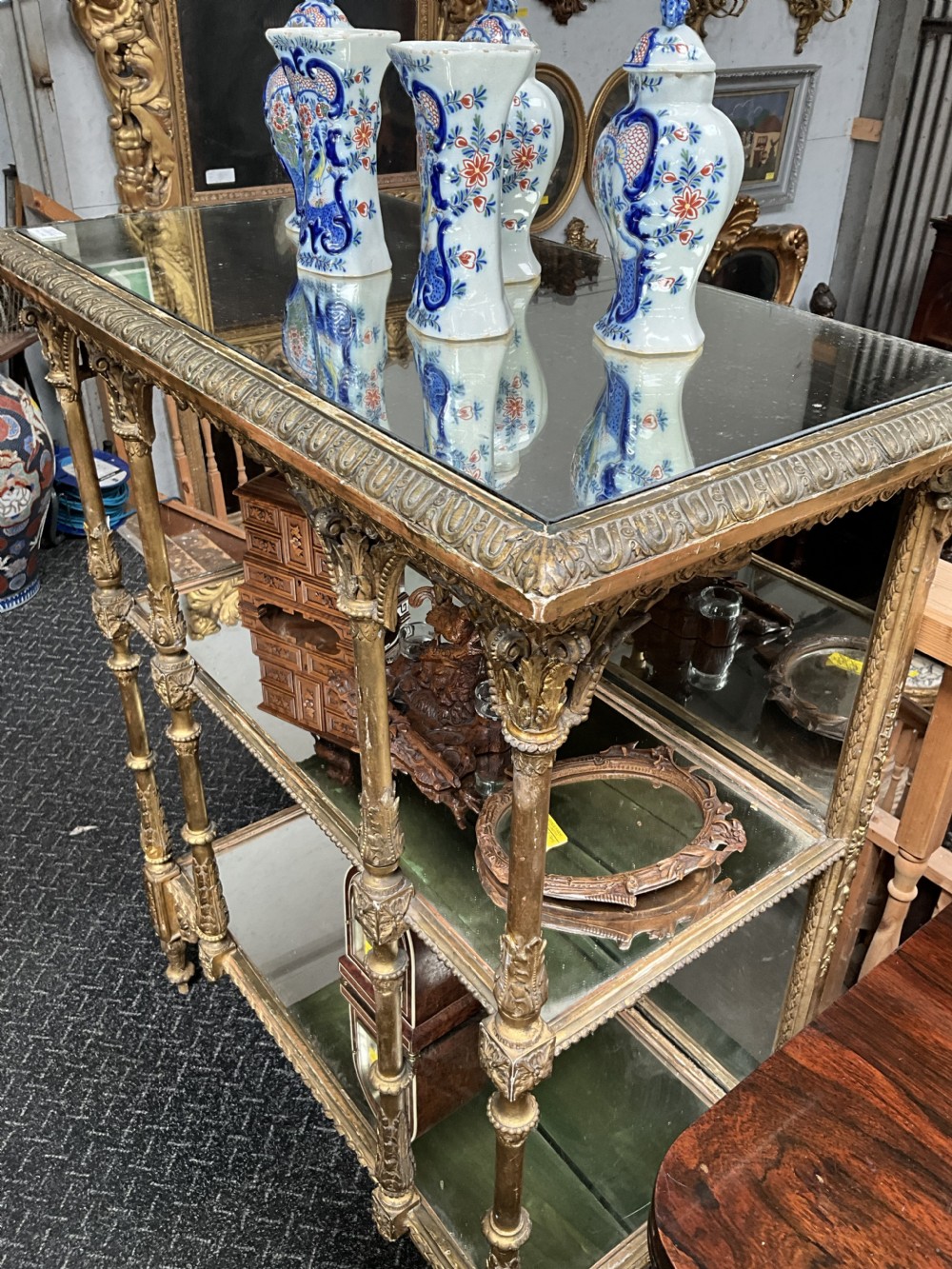 An ornate Victorian giltwood display shelf featuring three glass-tiered levels, a mirrored back panel, and intricate carved gesso columns with decorative moulding. View VI
