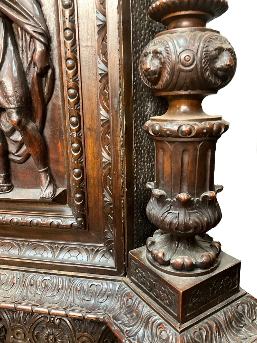A tall dark walnut cabinet featuring detailed classical relief carvings on two door panels flanked by turned columns. View VI