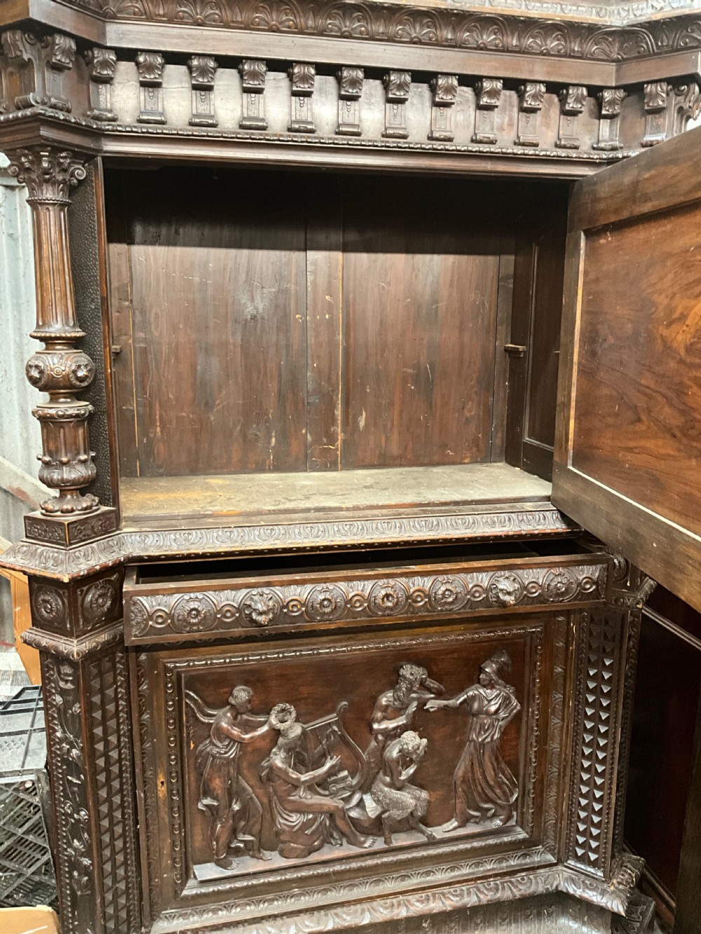 A tall dark walnut cabinet featuring detailed classical relief carvings on two door panels flanked by turned columns. View VII
