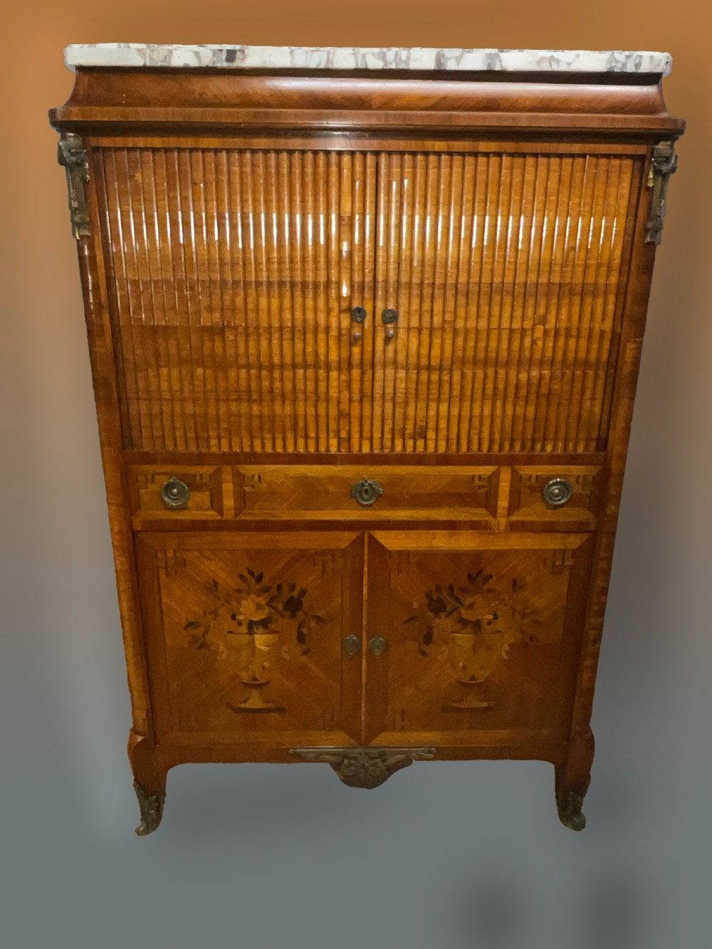 An antique French style marquetry writing cabinet featuring a marble top, tambour shutter front, middle drawer, floral inlaid doors, and brass mounts.