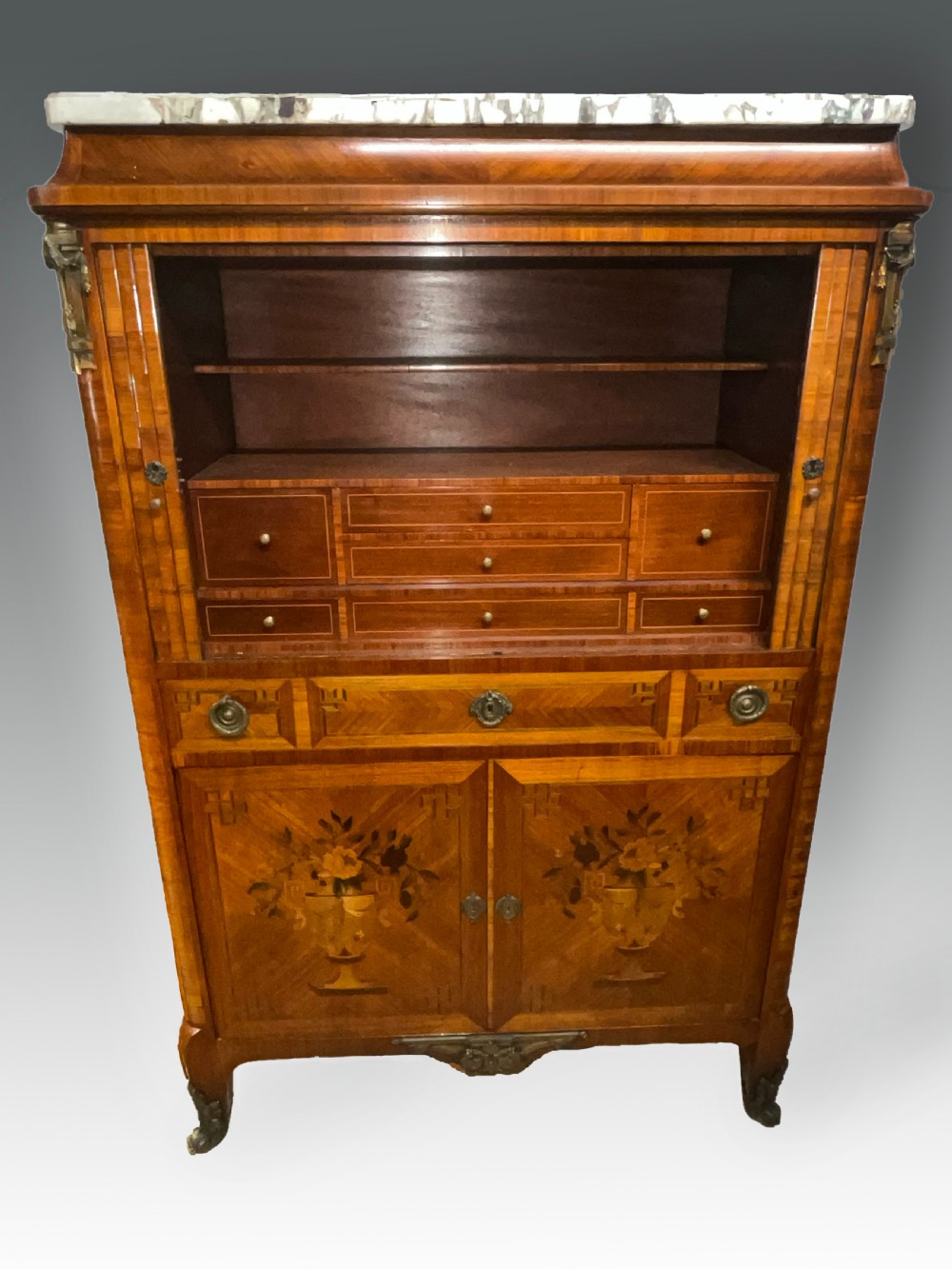 An antique French style marquetry writing cabinet featuring a marble top, tambour shutter front, middle drawer, floral inlaid doors, and brass mounts. View VI