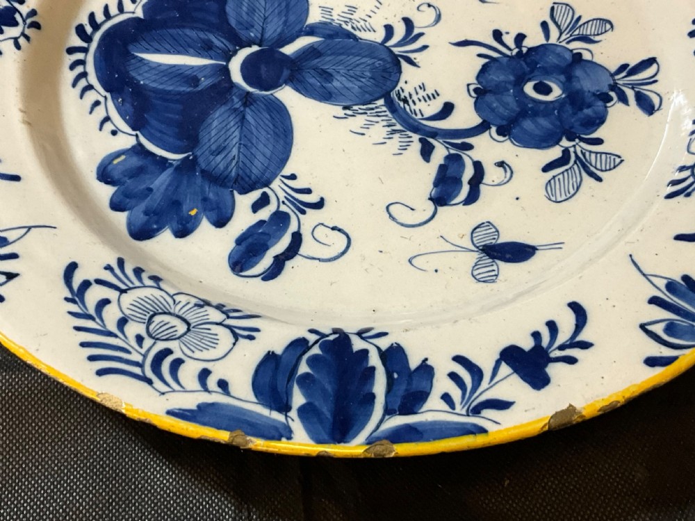 An antique circular tin-glazed earthenware plate featuring hand-painted cobalt blue floral motifs on a white background with a yellow rim. View V