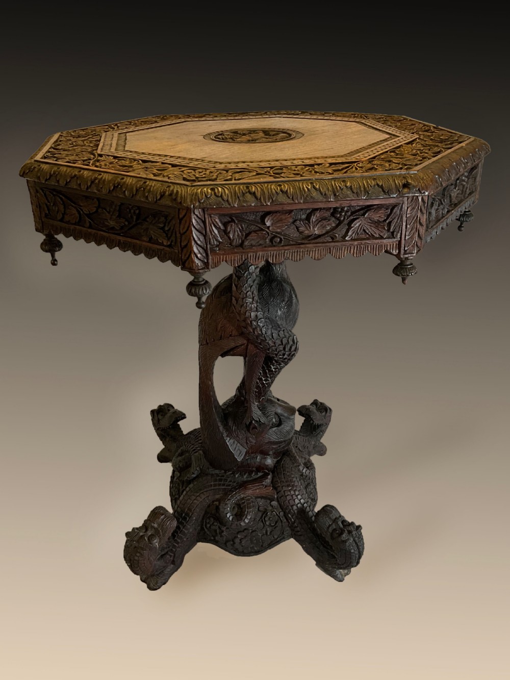 A 19th-century octagonal Burmese dark wood occasional table featuring detailed floral relief carvings, frieze drop finials, and a pedestal base carved as a mythical creature.
