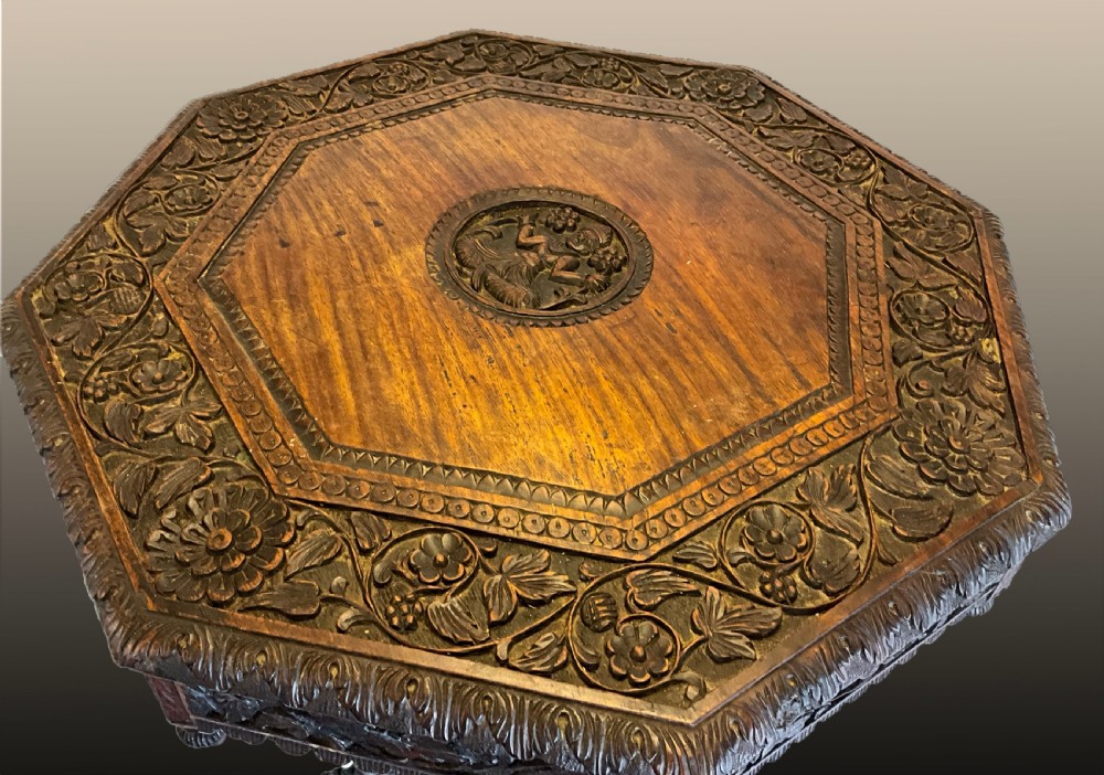 A 19th-century octagonal Burmese dark wood occasional table featuring detailed floral relief carvings, frieze drop finials, and a pedestal base carved as a mythical creature. View IV