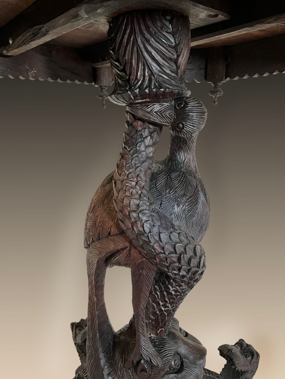 A 19th-century octagonal Burmese dark wood occasional table featuring detailed floral relief carvings, frieze drop finials, and a pedestal base carved as a mythical creature. View X