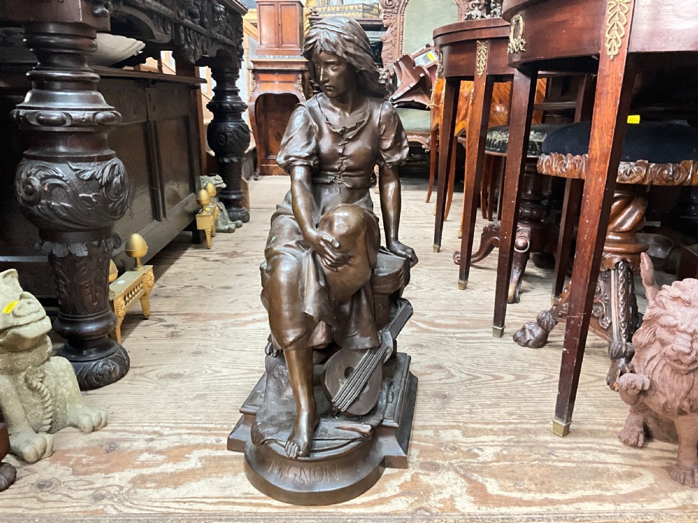 A dark patinated bronze statue depicting a seated young woman resting her foot on a rock next to a lute, with Mignon inscribed on the base.