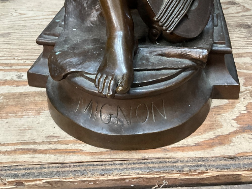 A dark patinated bronze statue depicting a seated young woman resting her foot on a rock next to a lute, with Mignon inscribed on the base. View III