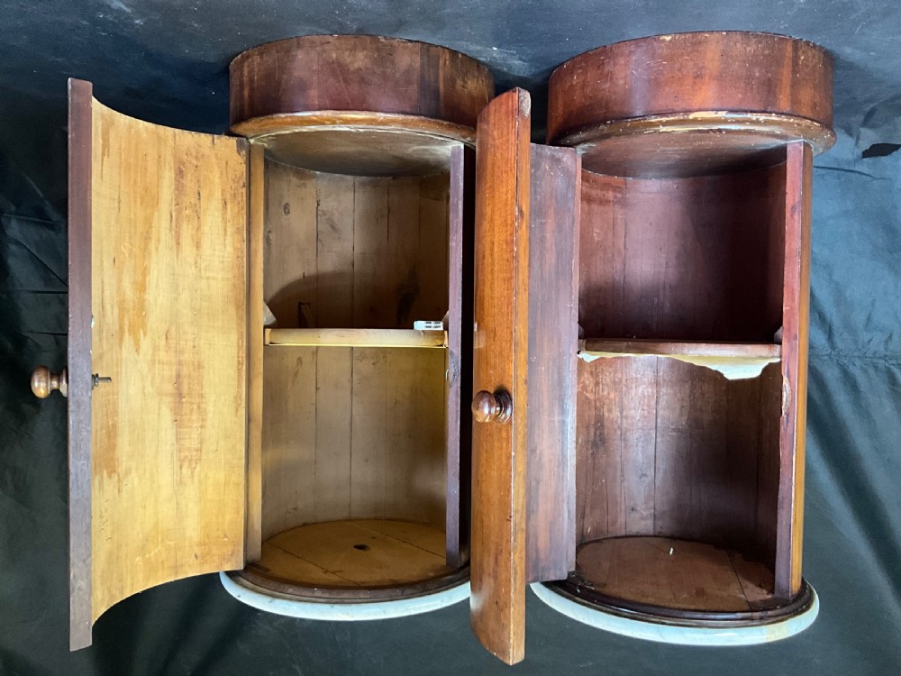 A matched pair of 19th-century Victorian cylindrical mahogany bedside cabinets featuring circular light grey marble tops, single curved doors with turned wooden knobs, and molded plinth bases. View II
