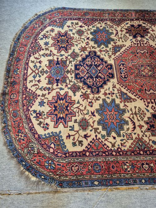 An antique hand-knotted Persian Bijar rug featuring a central terracotta medallion, geometric blue and red star motifs, a cream ground, and rounded curved ends. View III