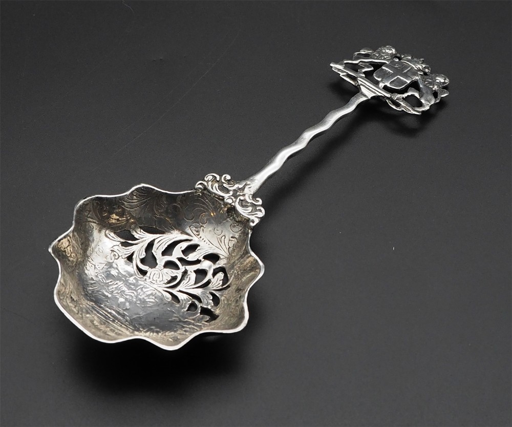 A Victorian sterling silver spoon featuring a pierced scalloped bowl, a wavy stem, and an armorial coat of arms finial.