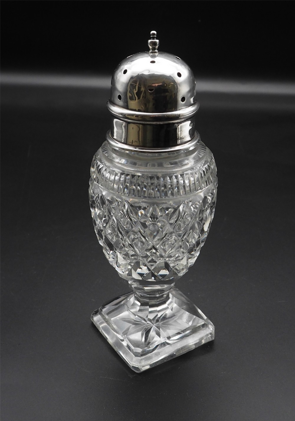 A clear cut glass sugar shaker featuring a diamond motif body, a square pedestal foot, and a perforated silver dome top.