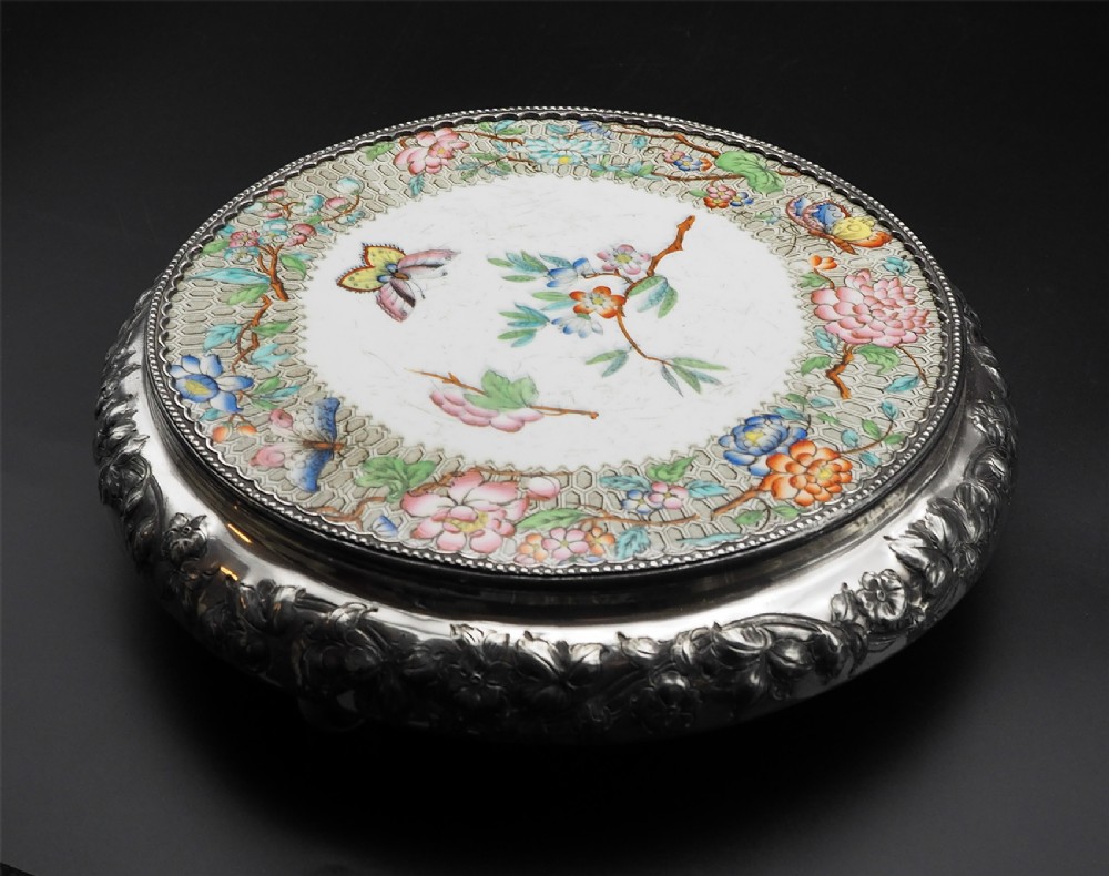 A circular Victorian ceramic table coaster featuring hand-painted floral and butterfly motifs on a white ground, framed by an embossed silver-plated rim.
