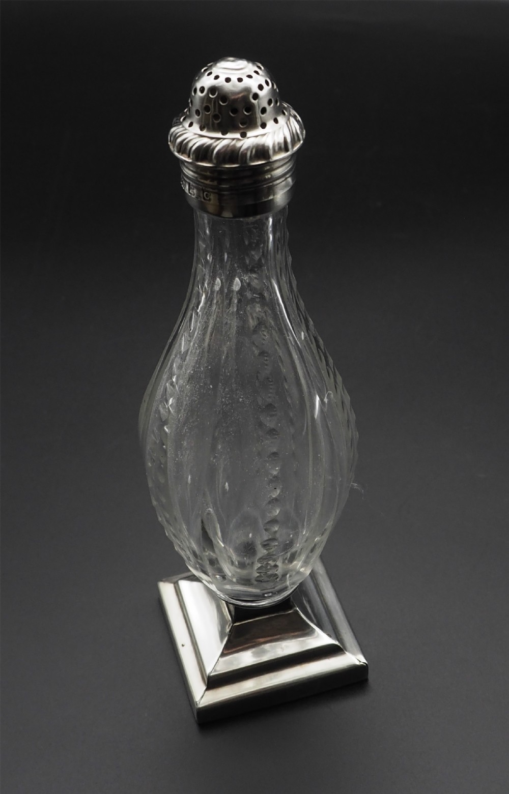 A Georgian cut glass sugar sifter featuring a faceted baluster glass body, square silver base, and hallmarked sterling silver pierced lid.