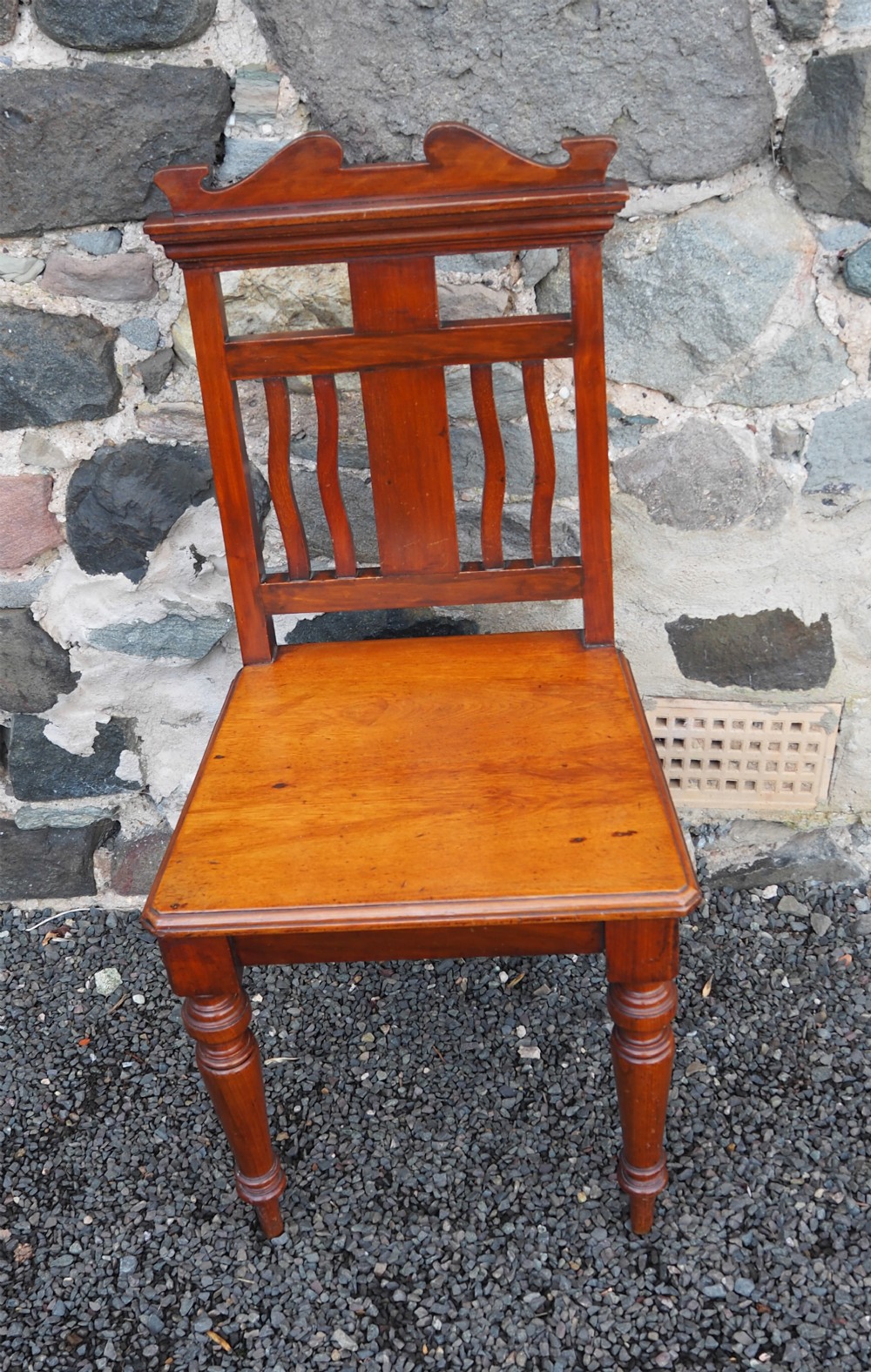 A late 19th-century American wooden hall chair featuring a shaped crest rail, vertical splat back, solid wooden seat, and turned front legs.