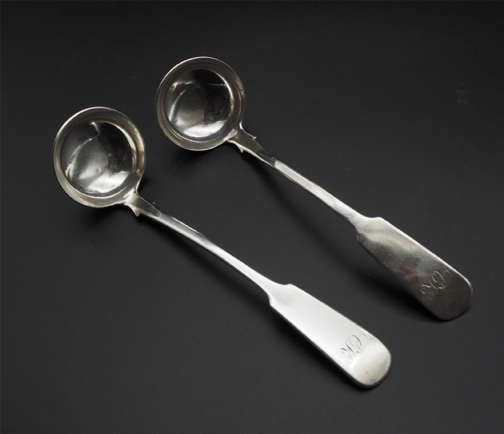 A pair of antique Scottish silver sauce ladles featuring circular bowls, flat fiddle handles, and engraved monograms displayed on a black surface.