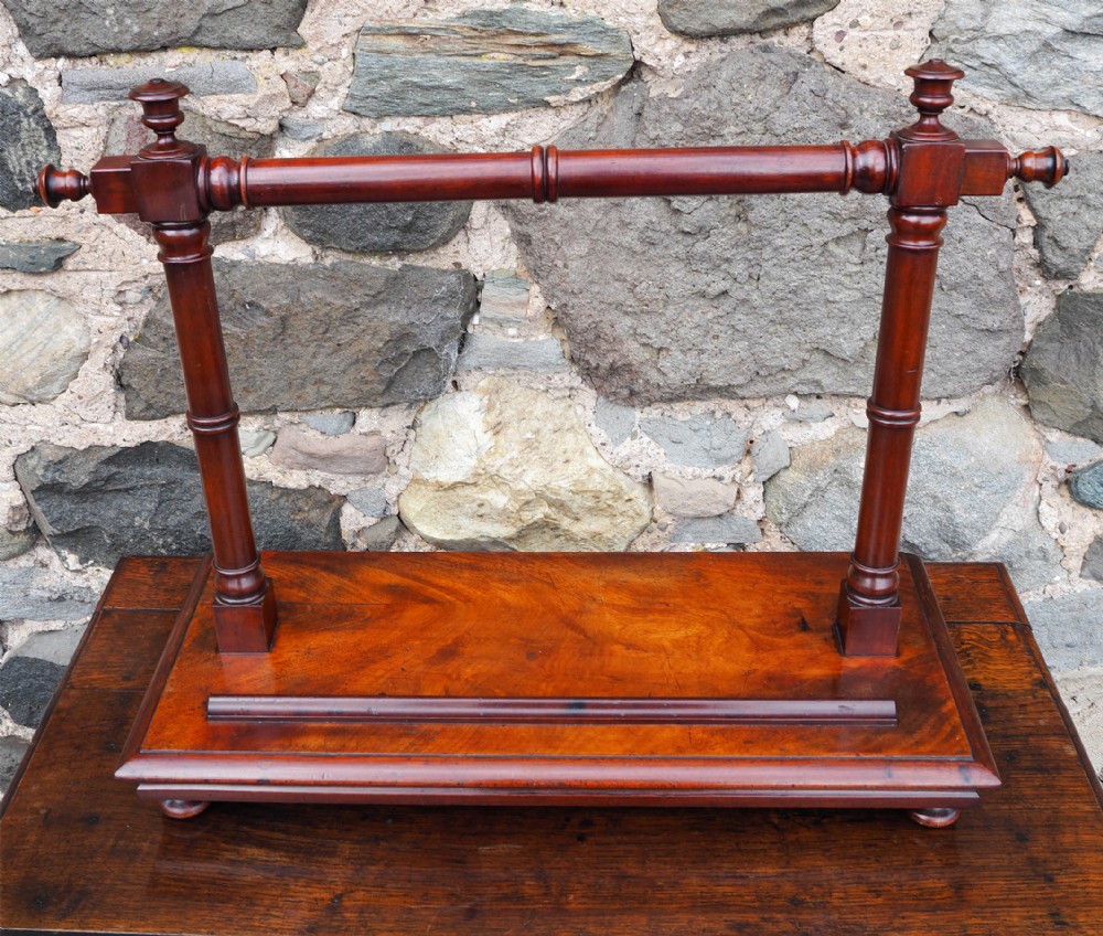 A dark brown mahogany tray stand featuring turned upright supports, a horizontal retaining bar, and a rectangular base with moulded edges.