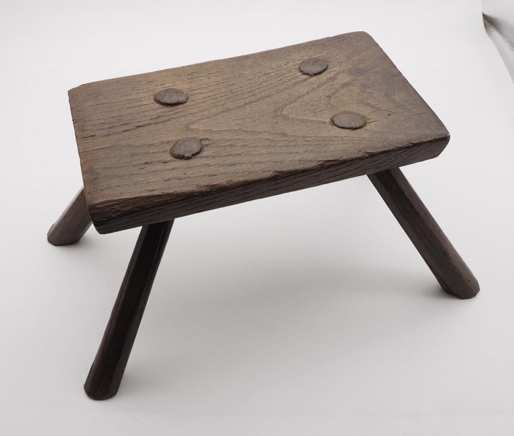 A low rectangular primitive oak stool featuring a thick hand-hewn seat and four round splayed legs visible through wedged tenons on top.