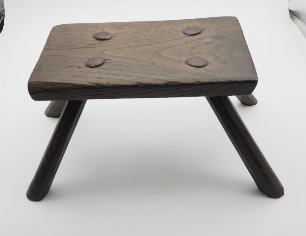 A low rectangular primitive oak stool featuring a thick hand-hewn seat and four round splayed legs visible through wedged tenons on top. View II