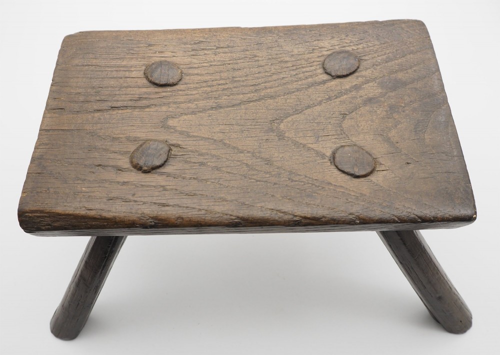 A low rectangular primitive oak stool featuring a thick hand-hewn seat and four round splayed legs visible through wedged tenons on top. View III