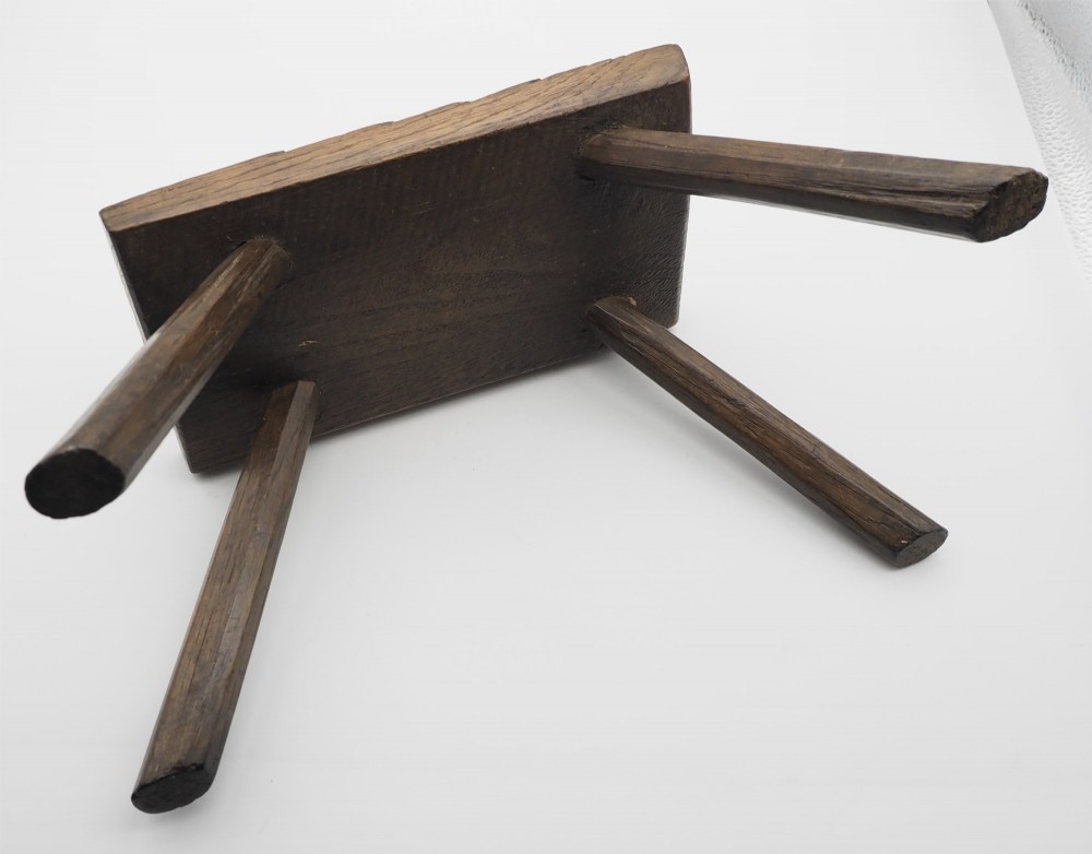 A low rectangular primitive oak stool featuring a thick hand-hewn seat and four round splayed legs visible through wedged tenons on top. View IV