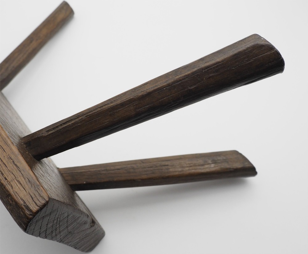 A low rectangular primitive oak stool featuring a thick hand-hewn seat and four round splayed legs visible through wedged tenons on top. View V