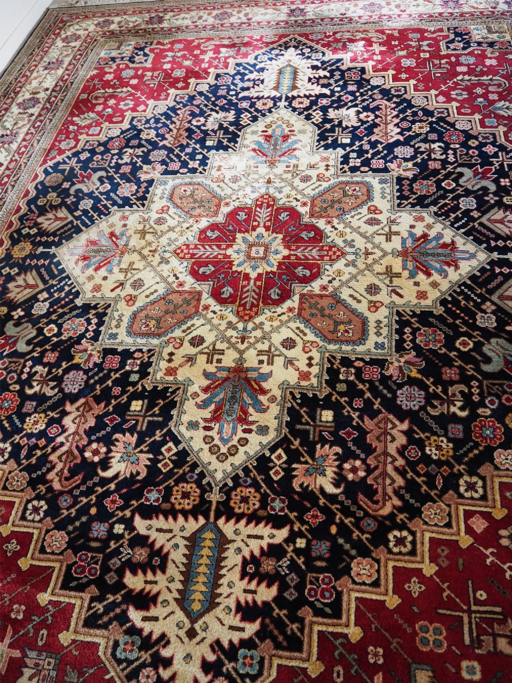 A large hand-knotted Persian wool carpet featuring a central cream medallion surrounded by navy blue and crimson sections filled with intricate floral motifs and framed by a detailed border. View V