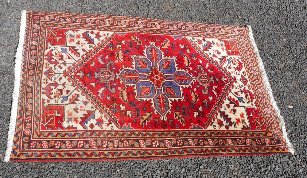 A small hand-knotted antique wool rug featuring a red central field with a blue star medallion, geometric cream spandrels, and a patterned border. View III