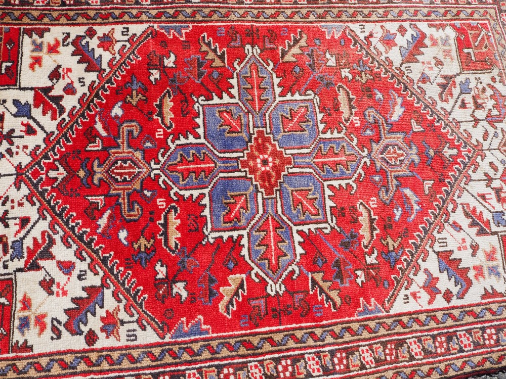 A small hand-knotted antique wool rug featuring a red central field with a blue star medallion, geometric cream spandrels, and a patterned border. View V