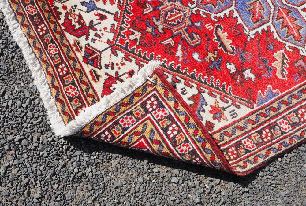 A small hand-knotted antique wool rug featuring a red central field with a blue star medallion, geometric cream spandrels, and a patterned border. View VI