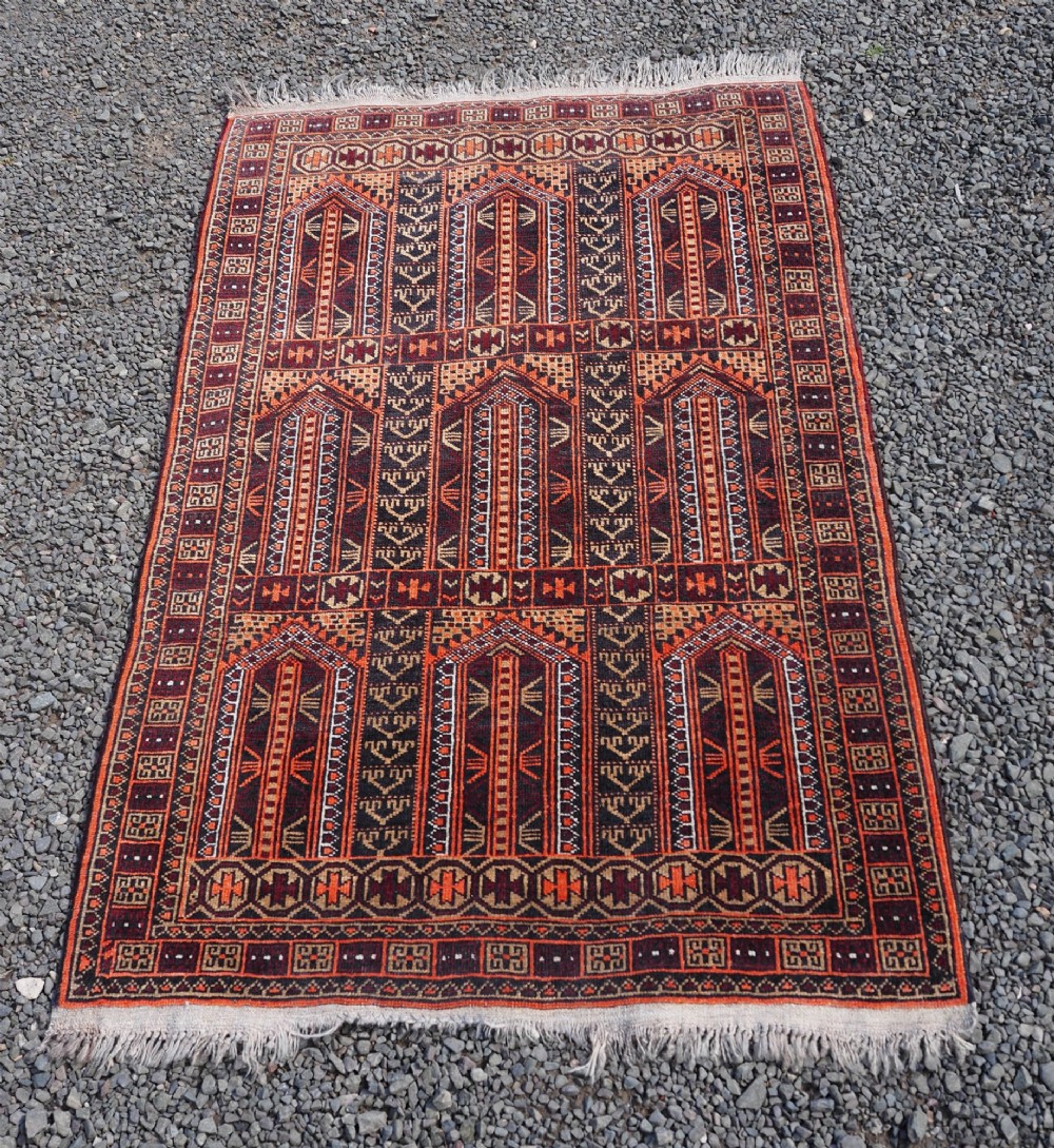 A rectangular hand-knotted wool Baluch rug featuring rows of geometric arched panels in terracotta red, rust orange, and dark navy with fringed ends.
