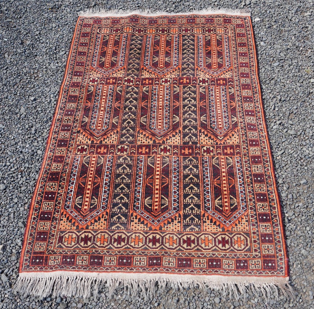 A rectangular hand-knotted wool Baluch rug featuring rows of geometric arched panels in terracotta red, rust orange, and dark navy with fringed ends. View II