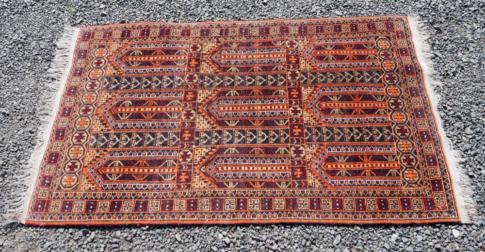 A rectangular hand-knotted wool Baluch rug featuring rows of geometric arched panels in terracotta red, rust orange, and dark navy with fringed ends. View III