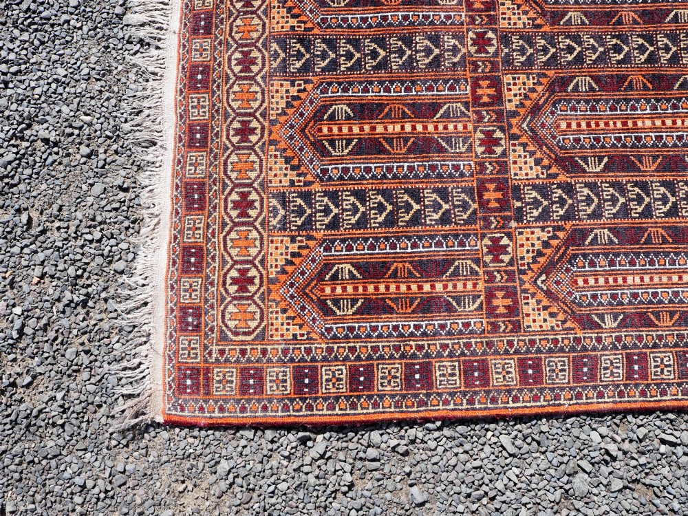A rectangular hand-knotted wool Baluch rug featuring rows of geometric arched panels in terracotta red, rust orange, and dark navy with fringed ends. View IV
