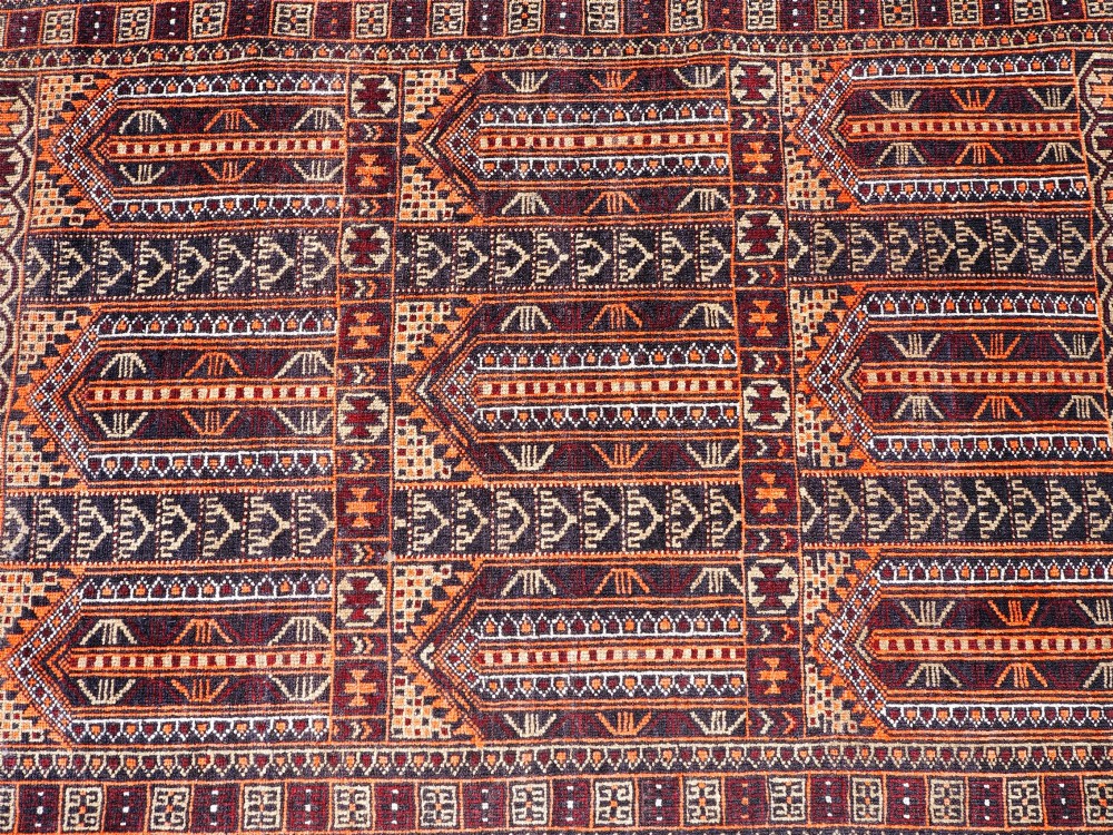 A rectangular hand-knotted wool Baluch rug featuring rows of geometric arched panels in terracotta red, rust orange, and dark navy with fringed ends. View V