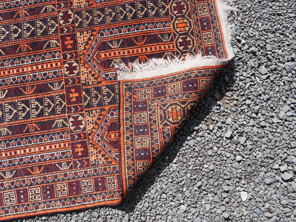 A rectangular hand-knotted wool Baluch rug featuring rows of geometric arched panels in terracotta red, rust orange, and dark navy with fringed ends. View VI