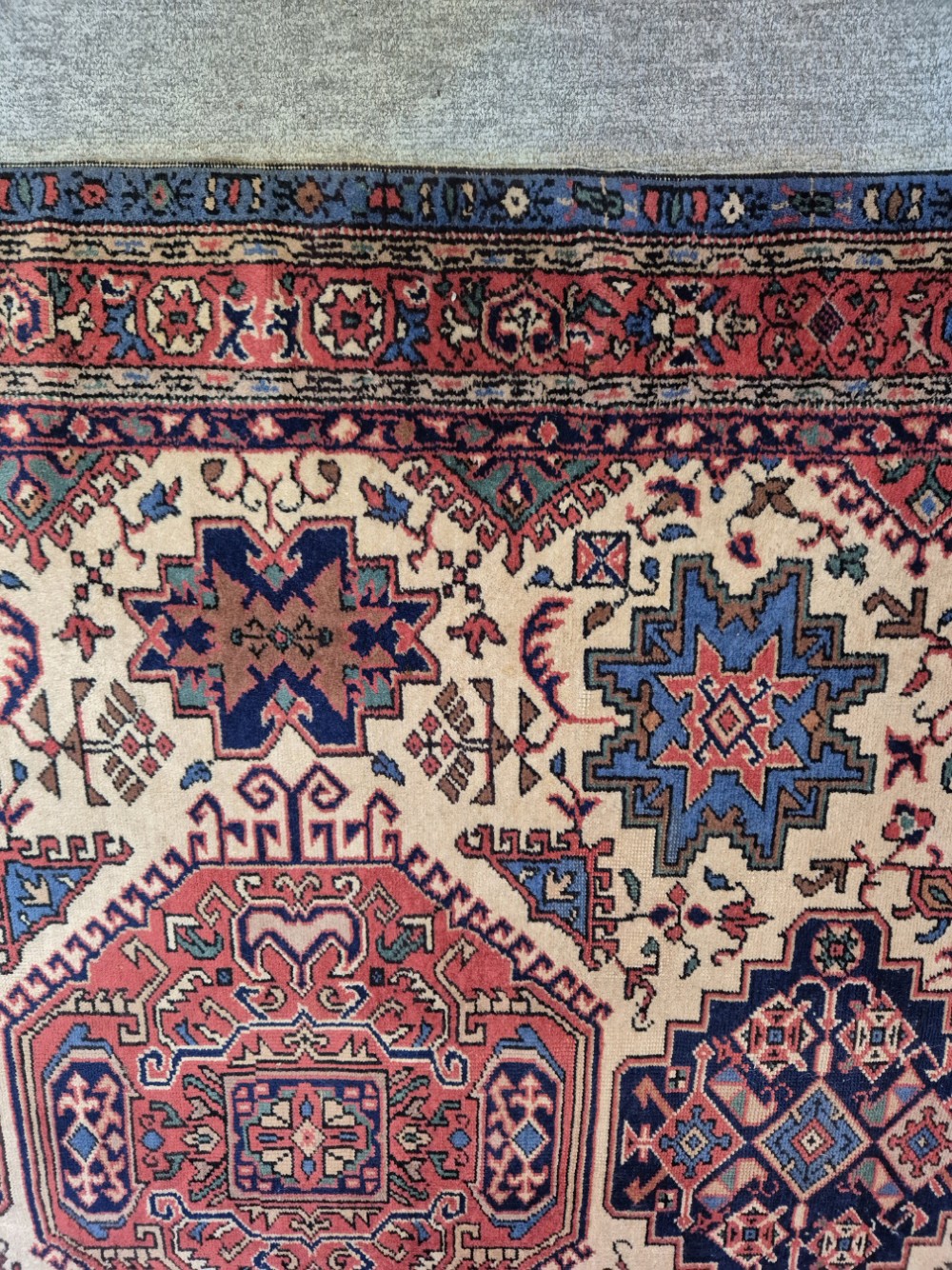An antique hand-knotted Persian Bijar rug featuring a central terracotta medallion, geometric blue and red star motifs, a cream ground, and rounded curved ends. View VII